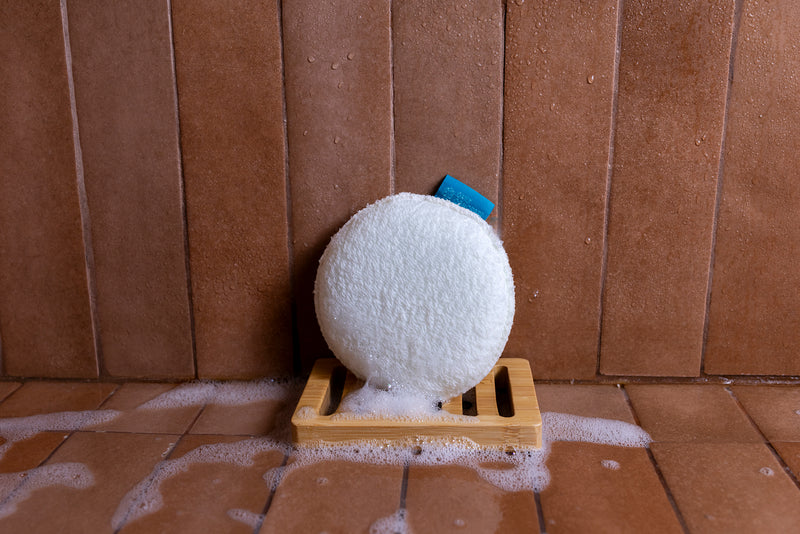 A white textured bath sponge resting on a small wooden soap dish with soap foam around it, set against a brown tiled wall.
