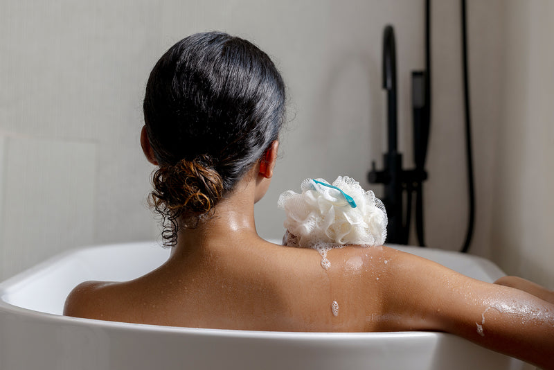 A woman with wet dark hair sits in a white bathtub, viewed from behind, with a white loofah sponge on her shoulder in a bathroom setting.