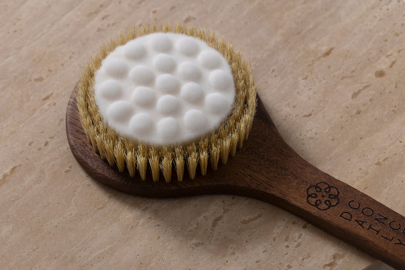 A close-up of a wooden body brush with natural bristles and a white textured foam pad, resting on a beige stone surface. The handle is engraved with the brand name Daily Concepts