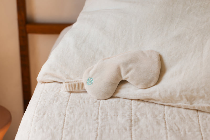 A cream-colored plush sleep mask with a small light blue embroidered logo, resting on a white quilted bedspread next to a white pillow.