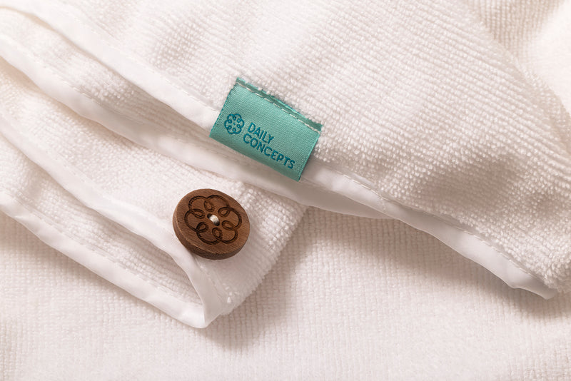 Close-up of a white textured hair towel with a turquoise fabric label reading 'DAILY CONCEPTS' and a small round brown wooden button with an engraved floral pattern.