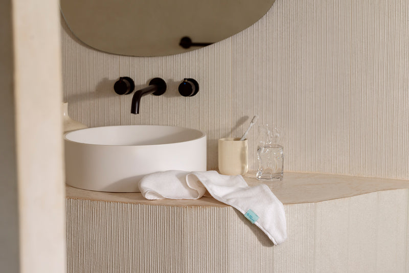 A modern bathroom vanity setup with a white round vessel sink, matte black faucet controls, a light wood countertop, a folded white towel, a small cream cup, and a clear glass bottle against a ribbed beige wall.