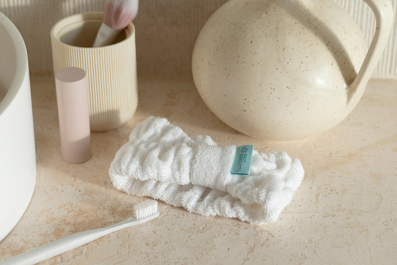 A folded white textured headband with a small blue-green brand tag lies on a beige stone surface, surrounded by bathroom-style props including a toothbrush, tooth holder, and ceramic vessel.