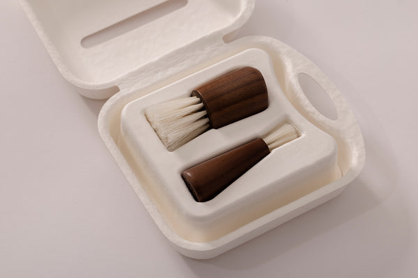 A two-piece wooden brush set with dark brown handles and off-white bristles, presented in a white molded sugar cane case on a light gray background.