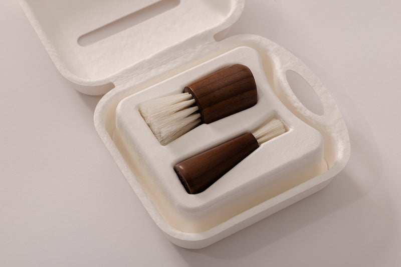 A two-piece wooden brush set with dark brown handles and off-white bristles, presented in a white molded sugar cane case on a light gray background.