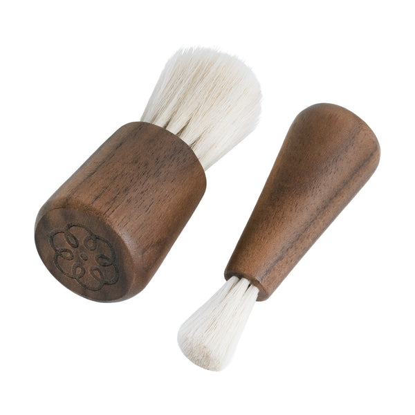 Two small wooden-handled brush tools with white bristles on a plain white background. One brush is shown from the end, revealing an engraved floral or clover-like pattern on the wooden cap, and the other is shown from the side.