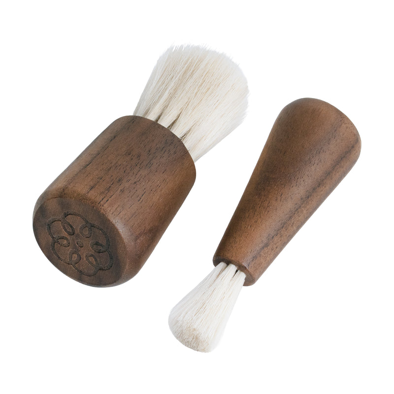 Two small wooden-handled brush tools with white bristles on a plain white background. One brush is shown from the end, revealing an engraved floral or clover-like pattern on the wooden cap, and the other is shown from the side.