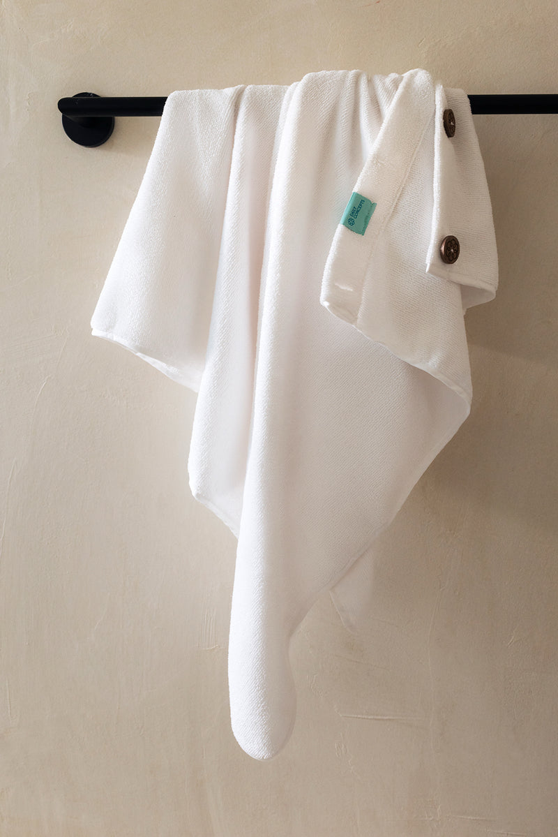 A white towel draped over a black metal rod against a textured beige wall, featuring a small turquoise tag and dark buttons on the corner.